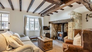 A cozy living room with exposed wooden beams, a stone fireplace, a sofa, a leather armchair, and a television. - 2 bedroom Cotswold Holiday Home - Abbots Cottage (Winchcombe)