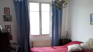 Un dormitorio con una cama grande, un radiador, una ventana con cortinas, un candelabro y una mesita de noche con lámpara.