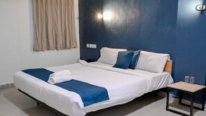 Una cama doble con ropa de cama blanca y azul, una mesita de noche pequeña con dos vasos, y una lámpara fijada a la pared.