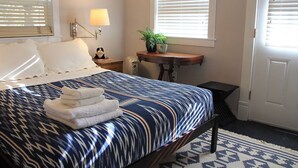 A neatly made bed with a patterned bedspread and folded towels, a bedside lamp, a small table with a plant, and a window with blinds. - Private, cozy, airy garden studio suite, for 1 or 2 guests (Portland)
