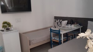 Una habitación pequeña y funcional con cama, escritorio con silla, televisor montado en la pared y un refrigerador pequeño.