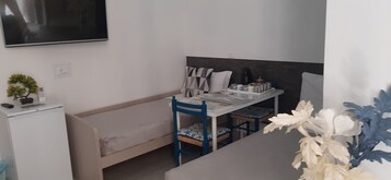 Una habitación pequeña y funcional con cama, escritorio con silla, televisor montado en la pared y un refrigerador pequeño.