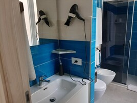Un baño con lavamanos blanco, espejo, secador de pelo montado en la pared, zona de ducha y inodoro.