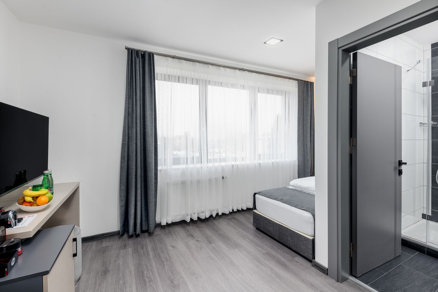 Habitación de hotel moderna con una cama, un escritorio con espejo, un televisor y una zona de ducha.