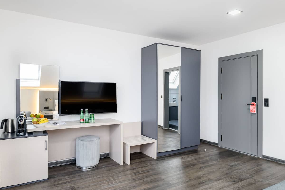 A modern hotel room with a flat-screen TV, a small kitchenette, a mirror, and a door with a red emergency exit sign.