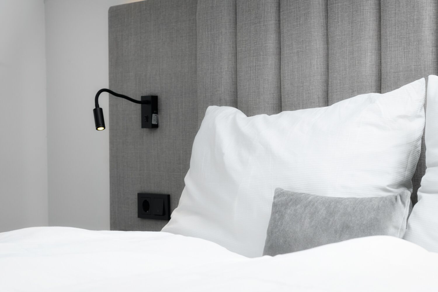 Ein modernes Hotelzimmer mit einem ordentlich bezogenen Bett, einer Wandleseleuchte und einem grauen Vorhang.