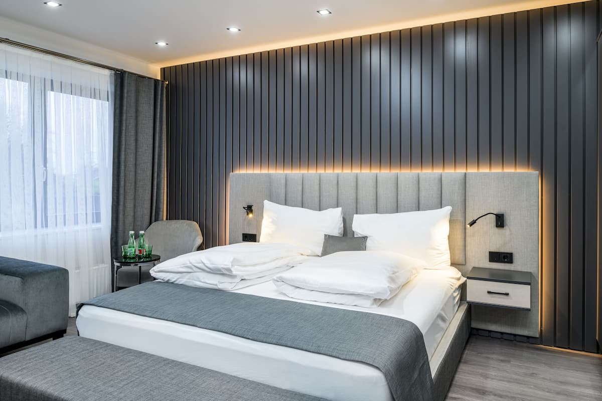 A modern hotel room with a large bed, a bedside table with a lamp, and a view of the outdoors through sheer curtains.