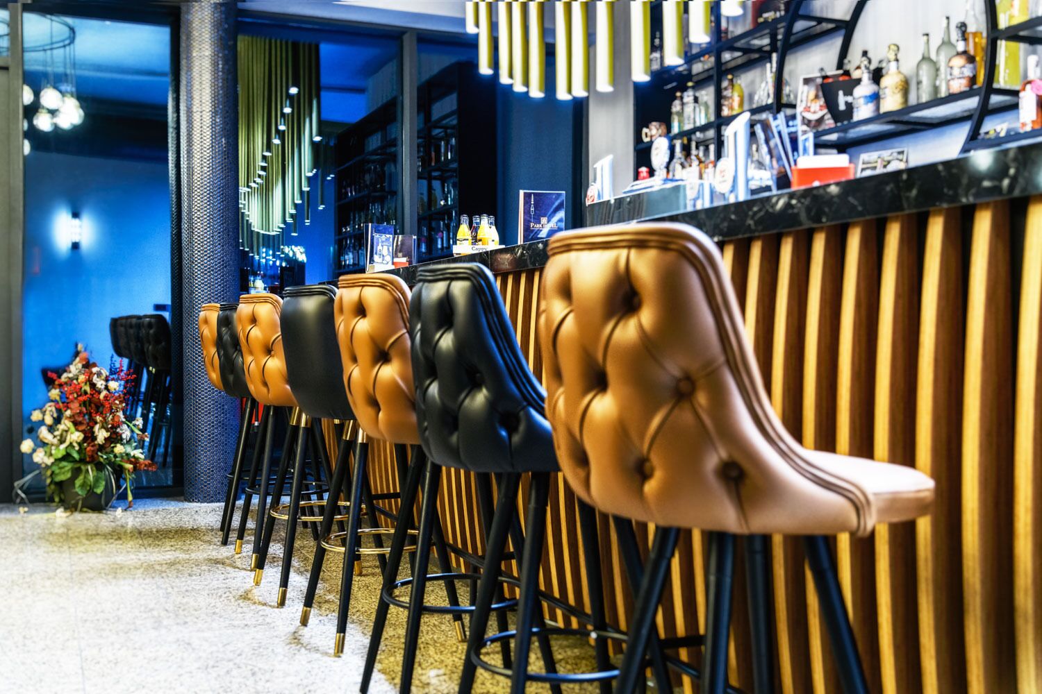 Eine Reihe von Barhocker mit gestopften Ledersitzen und schwarzen Metallbeinen, vor einer Bar mit einer Holzpaneelenwand und einer blauen Wand im Hintergrund.