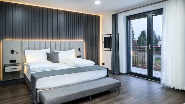A modern bedroom with a large bed, a nightstand, a TV mounted on the wall, and a sliding glass door to the outside. - Parkhotel Braunau  (Braunau am Inn)
