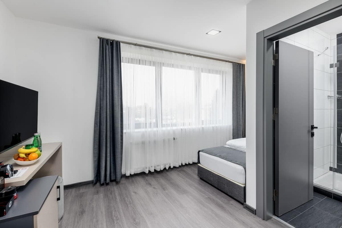 A modern hotel room with a bed, a desk with a mirror, a TV, and a shower area.