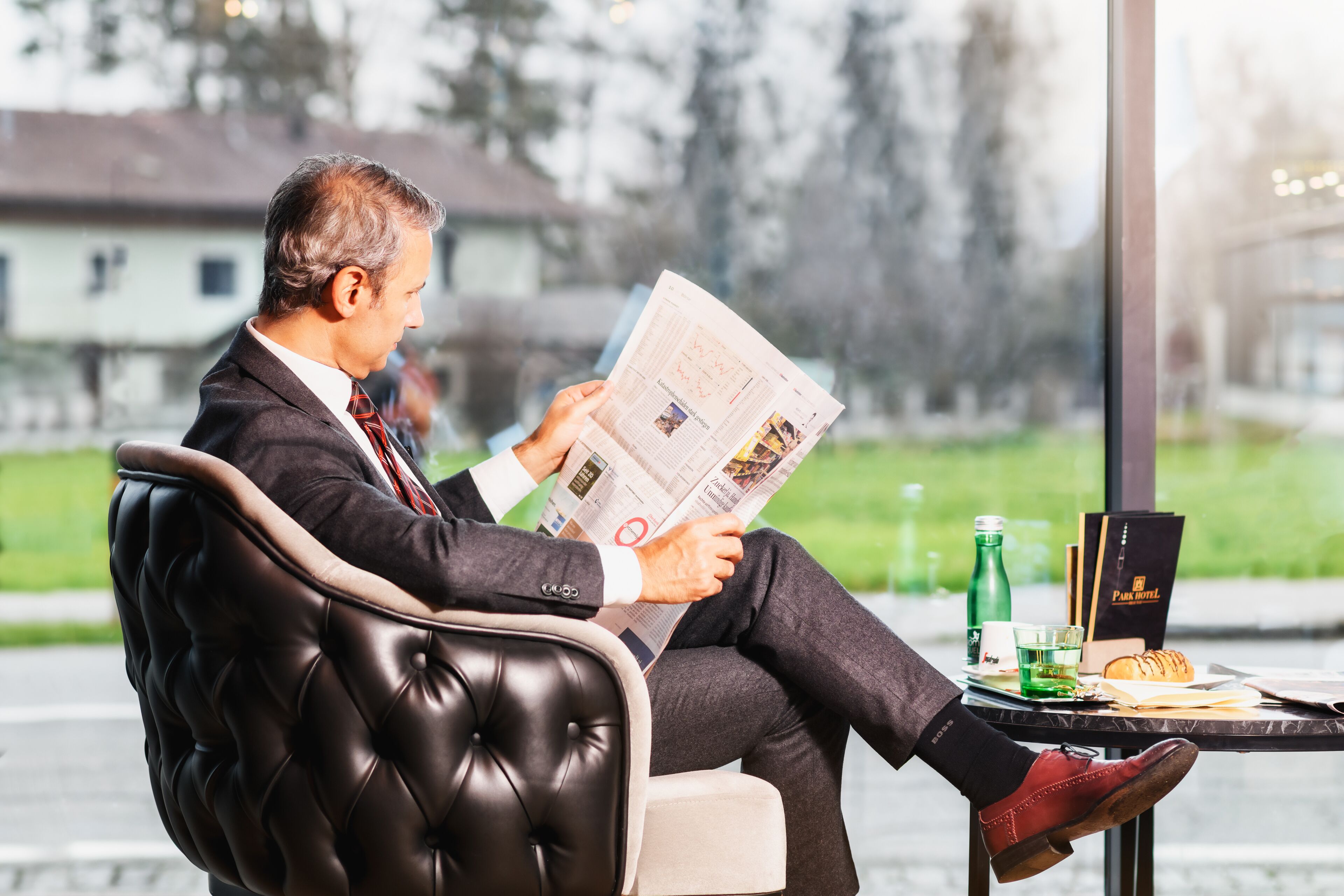 A man reading a newspaper in a leather chair with a table beside him containing a bottle, glasses, and a menu.