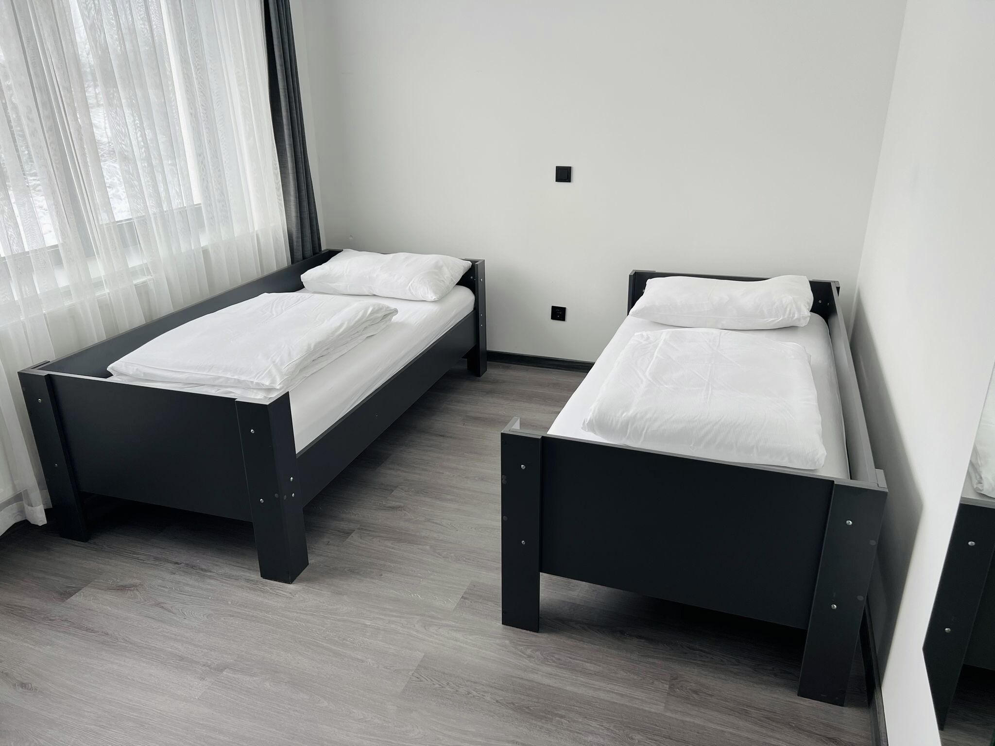 Ein Zimmer mit zwei Einzelbetten, einem Fenster mit leichtem Vorhang und einem hellen Holzboden.