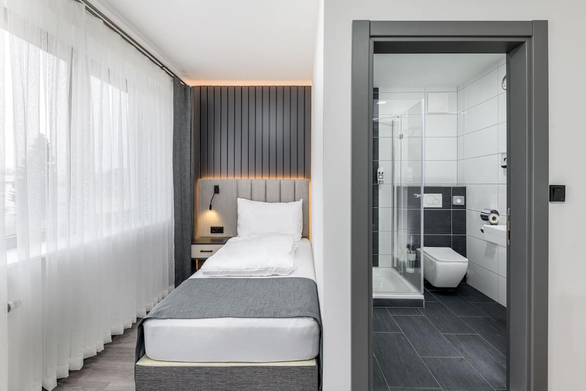 A modern hotel room with a bed, bedside table, and a view of the city through sheer curtains.