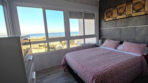 Quarto com uma grande janela, cama com roupa de cama estampada, cabeceira com painel decorativo e cômoda.