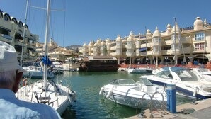 Uma marina com barcos atracados, um homem com chapéu branco e edifícios com arquitetura tradicional.