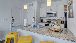 A kitchen with white cabinets, a granite countertop, a stainless steel microwave, a stove, and a sink. There are two yellow stools and a decorative tray with glassware. - PrimRose Retreat in Orange Pk Nr. NAS JAX& ENTMT. (Jacksonville)