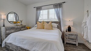 A bedroom with a wooden bed frame, white bedding, a bedside table with a lamp, a mirror, and a window with curtains. - PrimRose Retreat in Orange Pk Nr. NAS JAX& ENTMT. (Jacksonville)