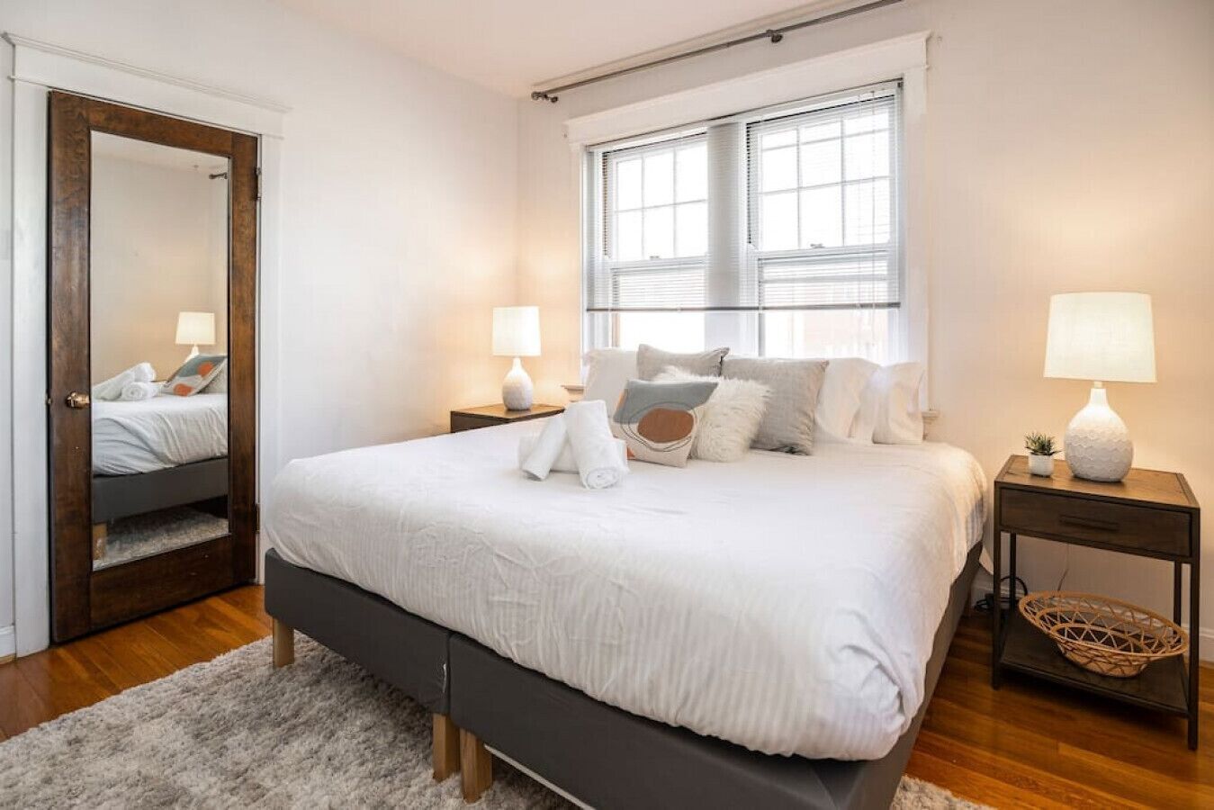 Un dormitorio ordenado con una cama grande, dos mesitas de noche con lámparas, un espejo y una ventana con persianas.
