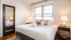 Een keurig opgemaakte slaapkamer met een groot bed, twee nachtkastjes met lampen, een spiegel en een raam met zonwering.