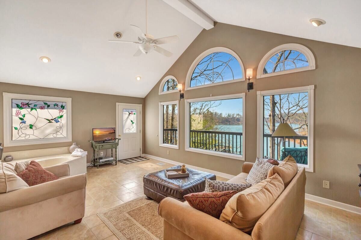 A spacious living room with a high ceiling, large windows, a ceiling fan, and a fireplace.