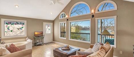 A spacious living room with a high ceiling, large windows, a ceiling fan, and a fireplace.