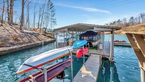 A dock with kayaks and a covered area by a calm water body, surrounded by trees. - Spacious 6BR Greene Lodge on Lake Keowee, perfect for family getaways! (Six Mile)