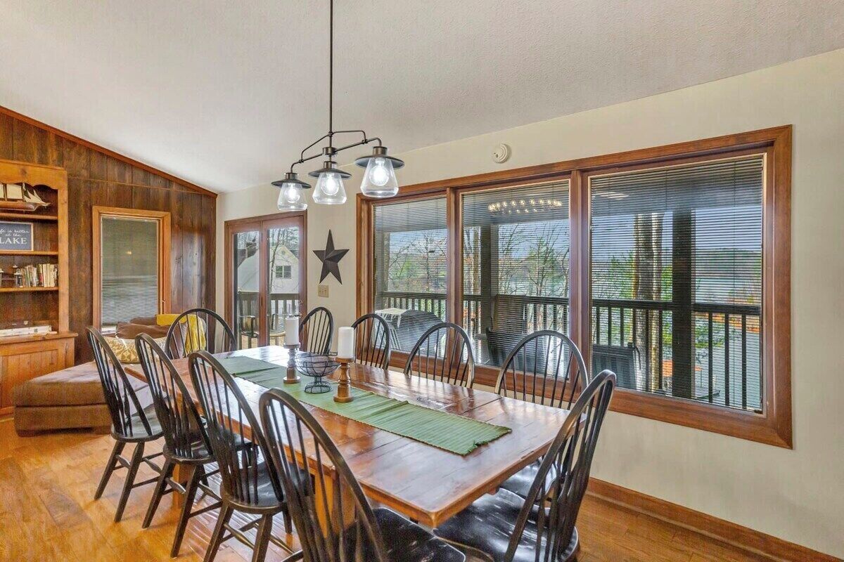A dining area with a wooden table, black chairs, and a chandelier. There are large windows with blinds and a star decoration on the wall.