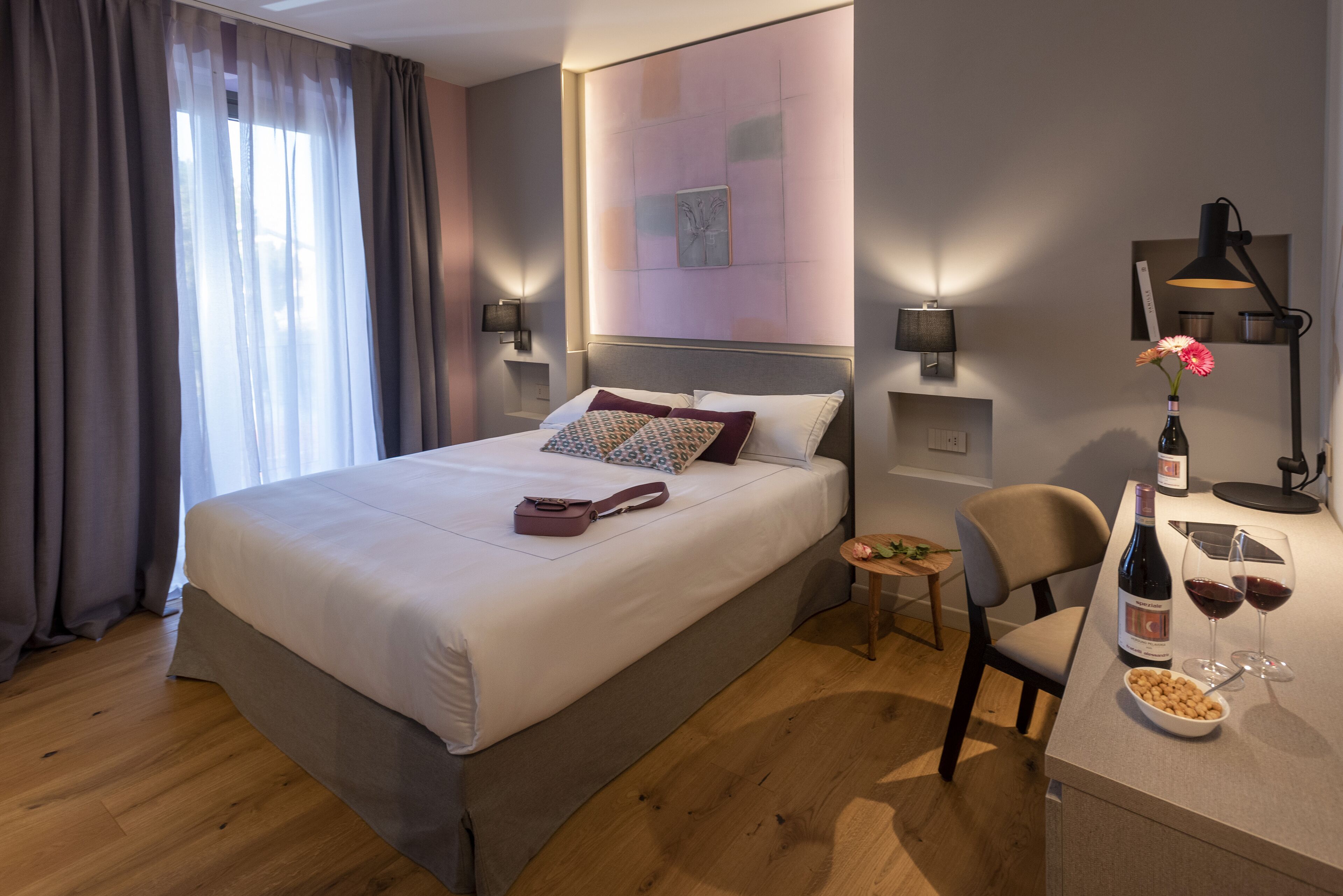 Habitación de hotel con una cama grande, mesitas de noche, un escritorio con una silla, una pequeña mesa con una botella y vasos, y una ventana con cortinas.