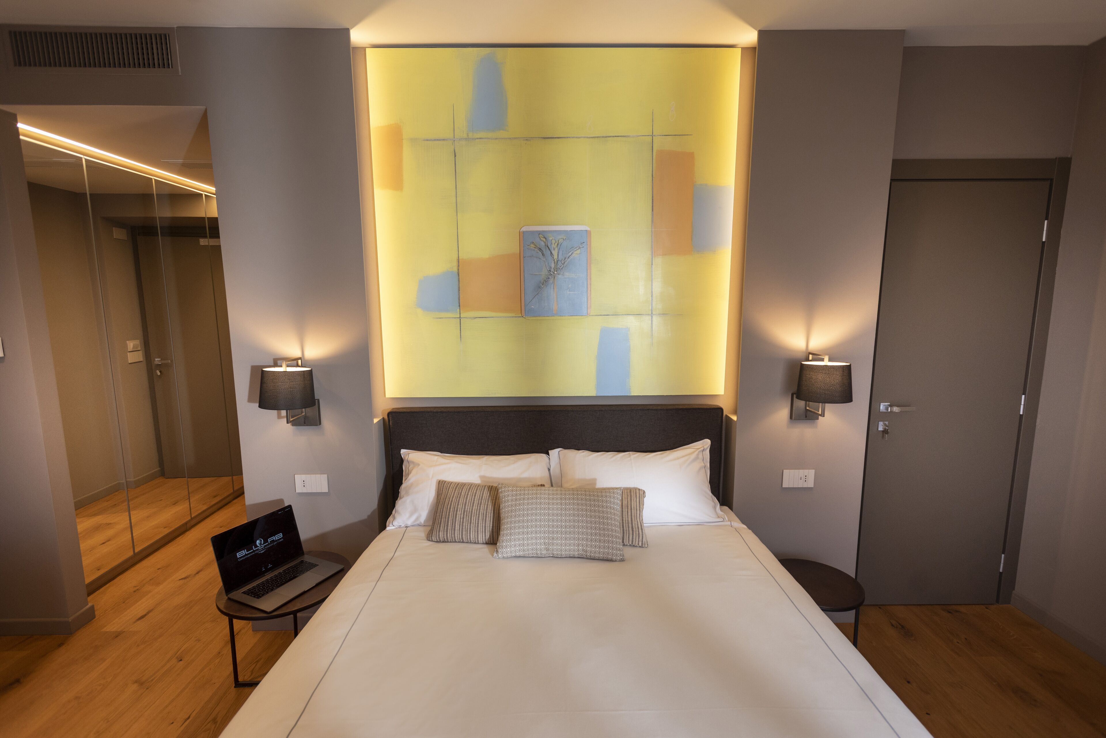 A modern hotel room with a large bed, a laptop on a side table, and a glass-enclosed shower area.