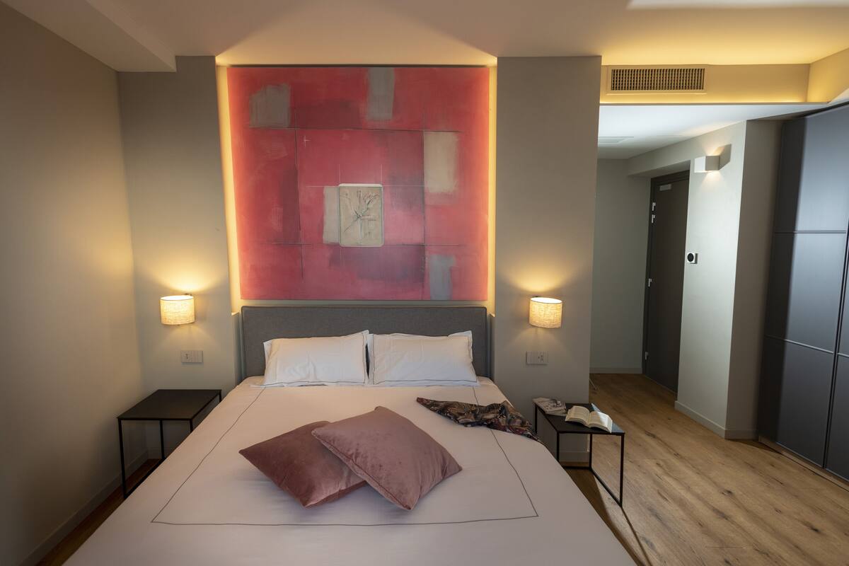Una habitación de hotel moderna con una cama grande, dos mesitas de noche, una mesilla auxiliar, un cuadro abstracto rojo en la pared y suelo de madera.