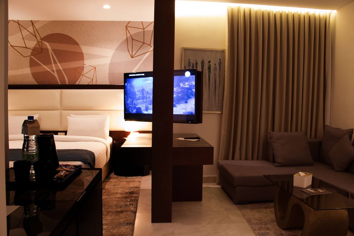 A modern hotel room with a bed, a sofa, a coffee table, and two televisions.