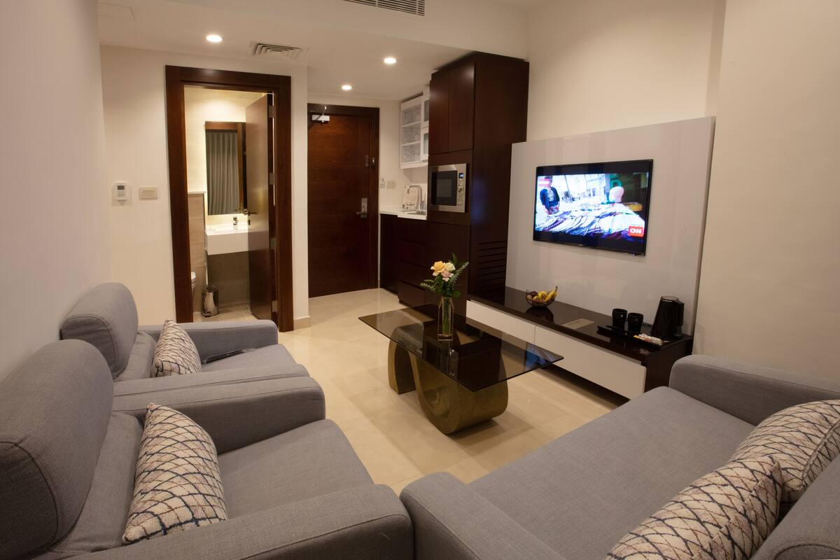 A modern living room with grey sofas, a glass coffee table, a flat-screen TV, and a microwave above a dark cabinet.