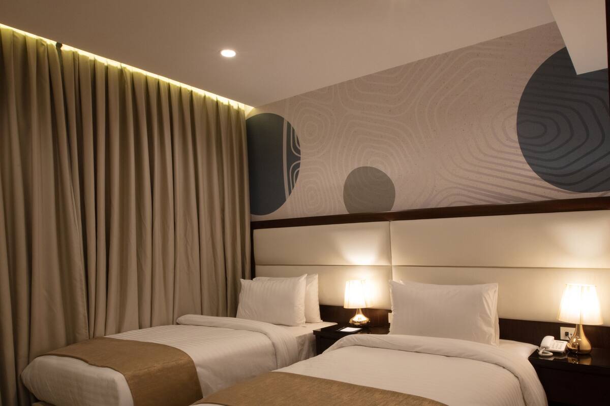 A hotel room with two beds, a large window with curtains, and a modern wall design.