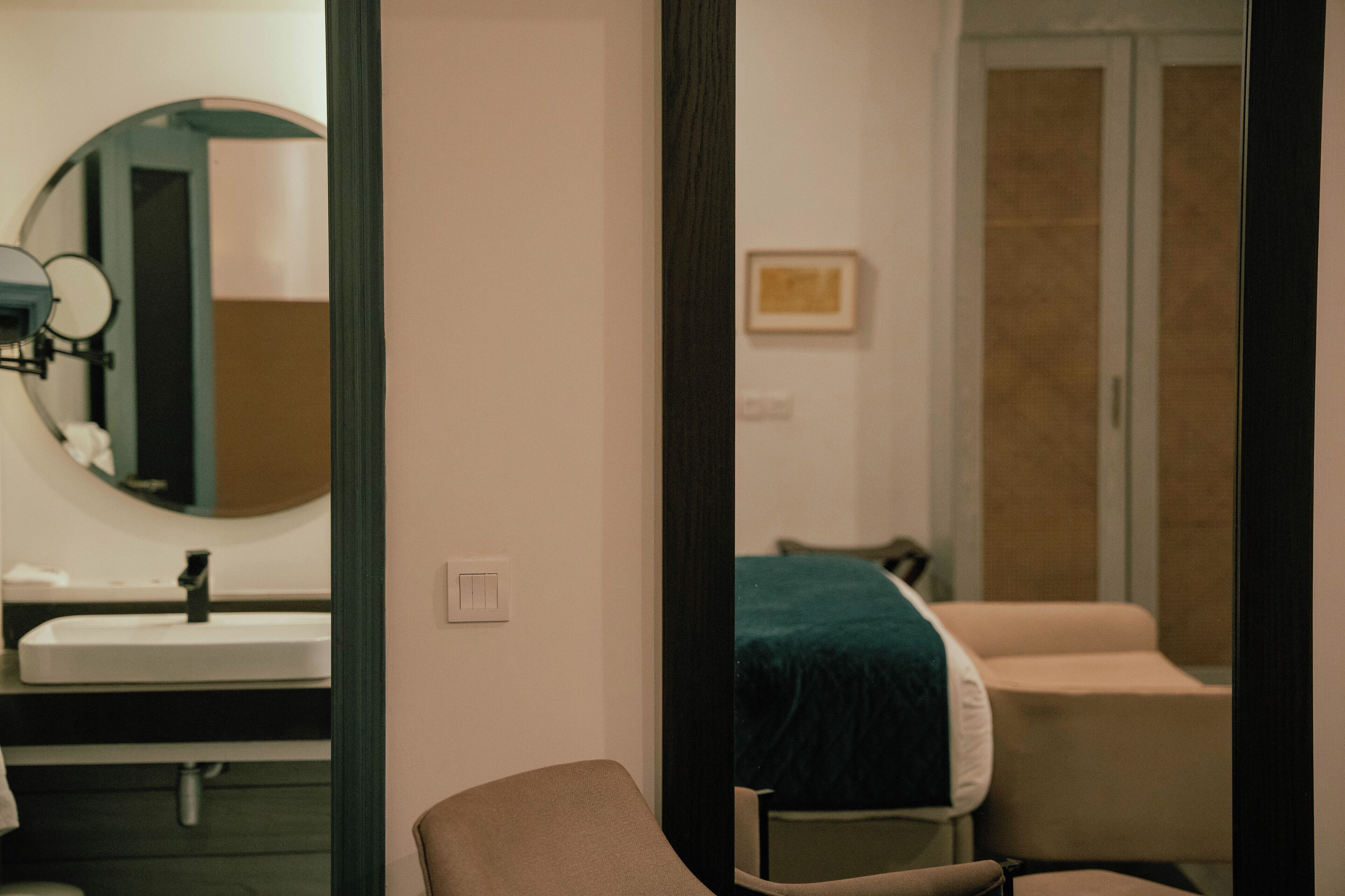 A room with a round mirror, a sink, a bed with a green blanket, and a chair.