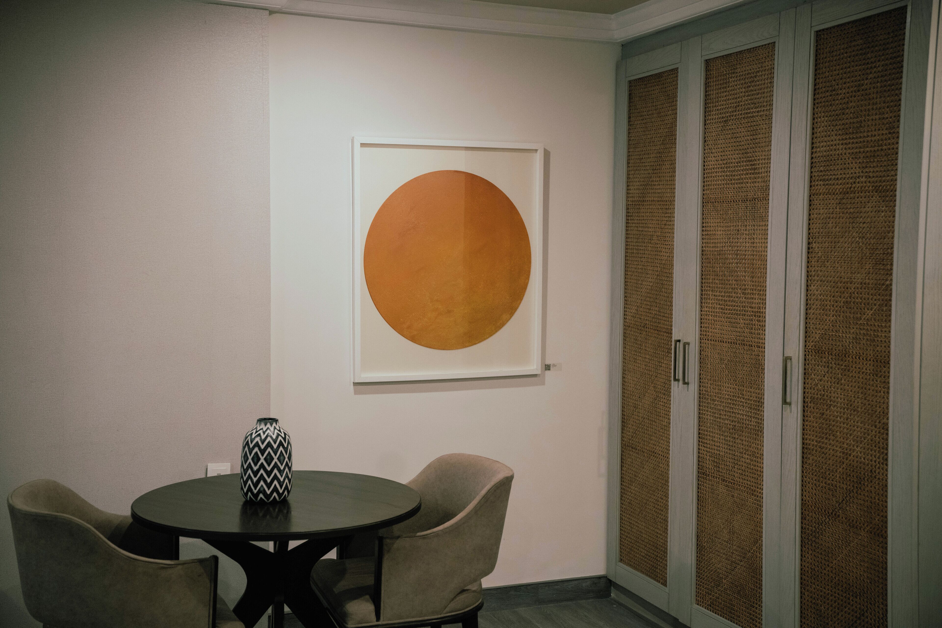 A room with a round table, two chairs, a framed artwork, and a door with a woven panel.
