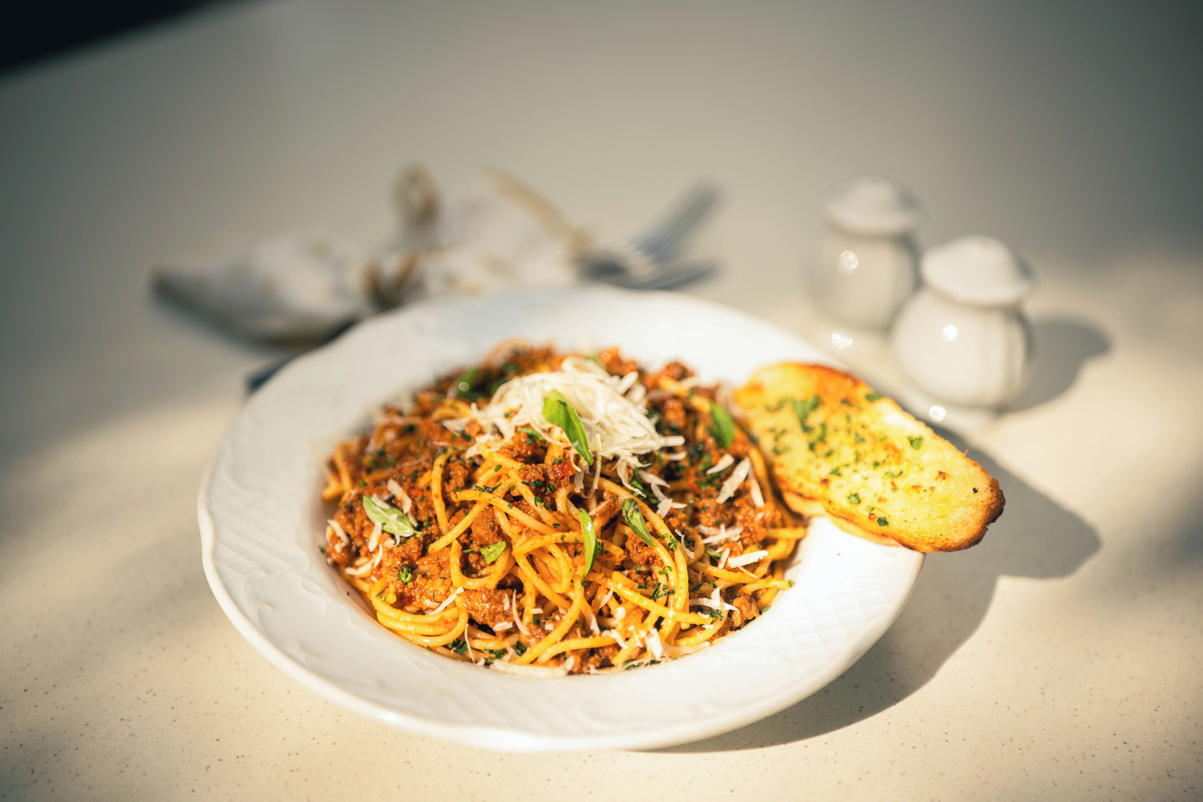 A plate of spaghetti with meat sauce, garnished with herbs, and a piece of toasted bread on a table.