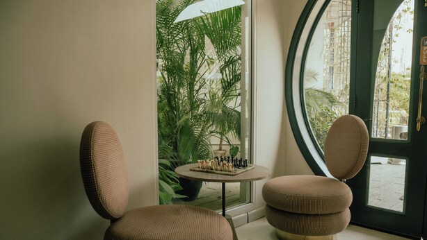 A room with two upholstered chairs, a small table with a chessboard, and a large window with a view of greenery and a building outside. -  Ambiance Boutique Art Hotel Karachi (Karachi)