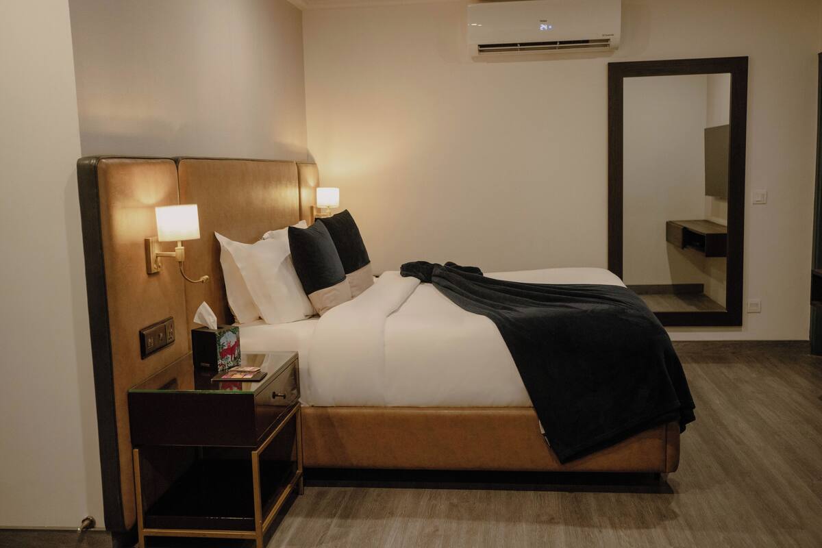 Una habitación de hotel con cama, mesita de noche, lámpara, equipo de aire acondicionado y un espejo.