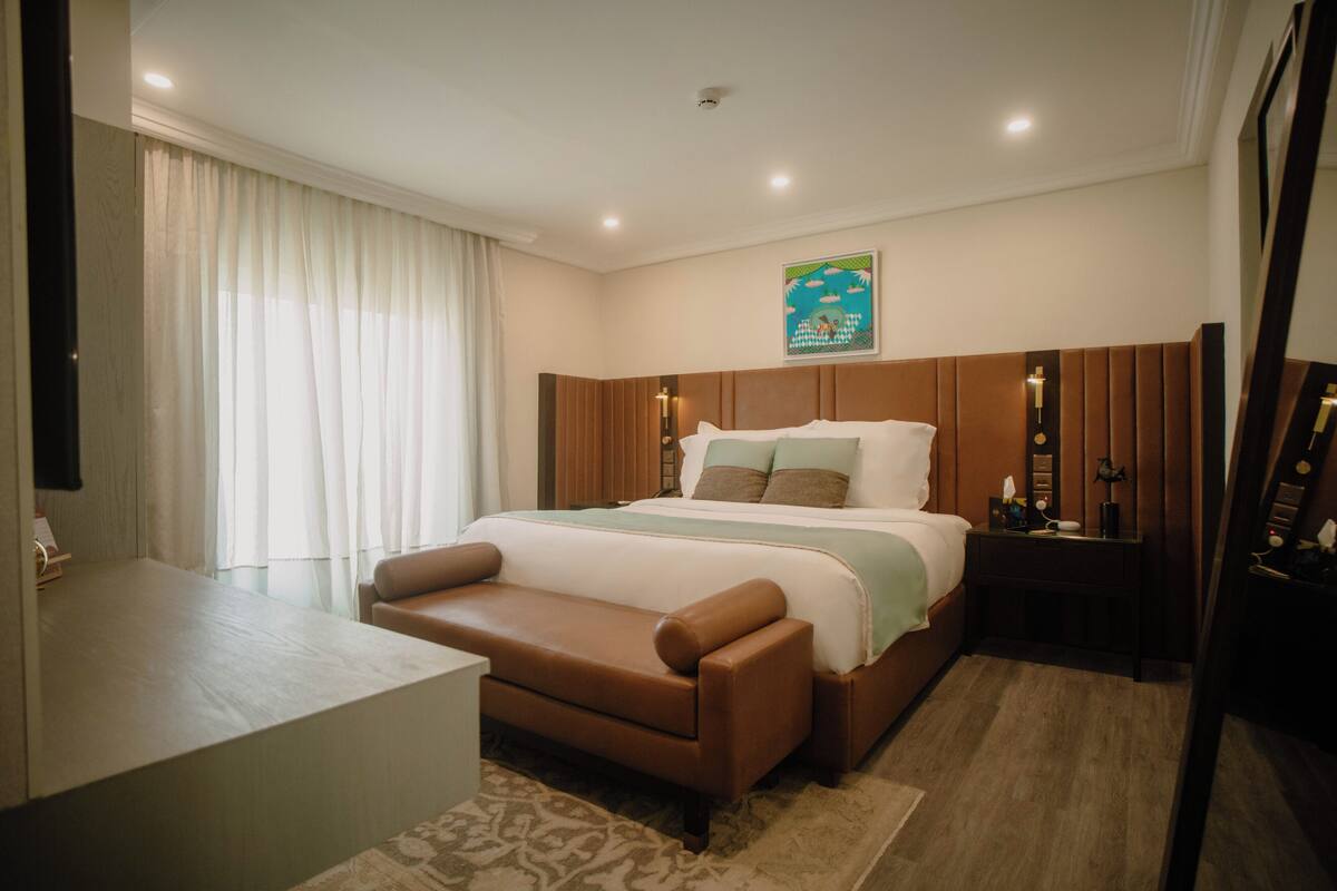 Un dormitorio de hotel con cama, mesitas de noche, un sofá y un ventanal con cortinas.