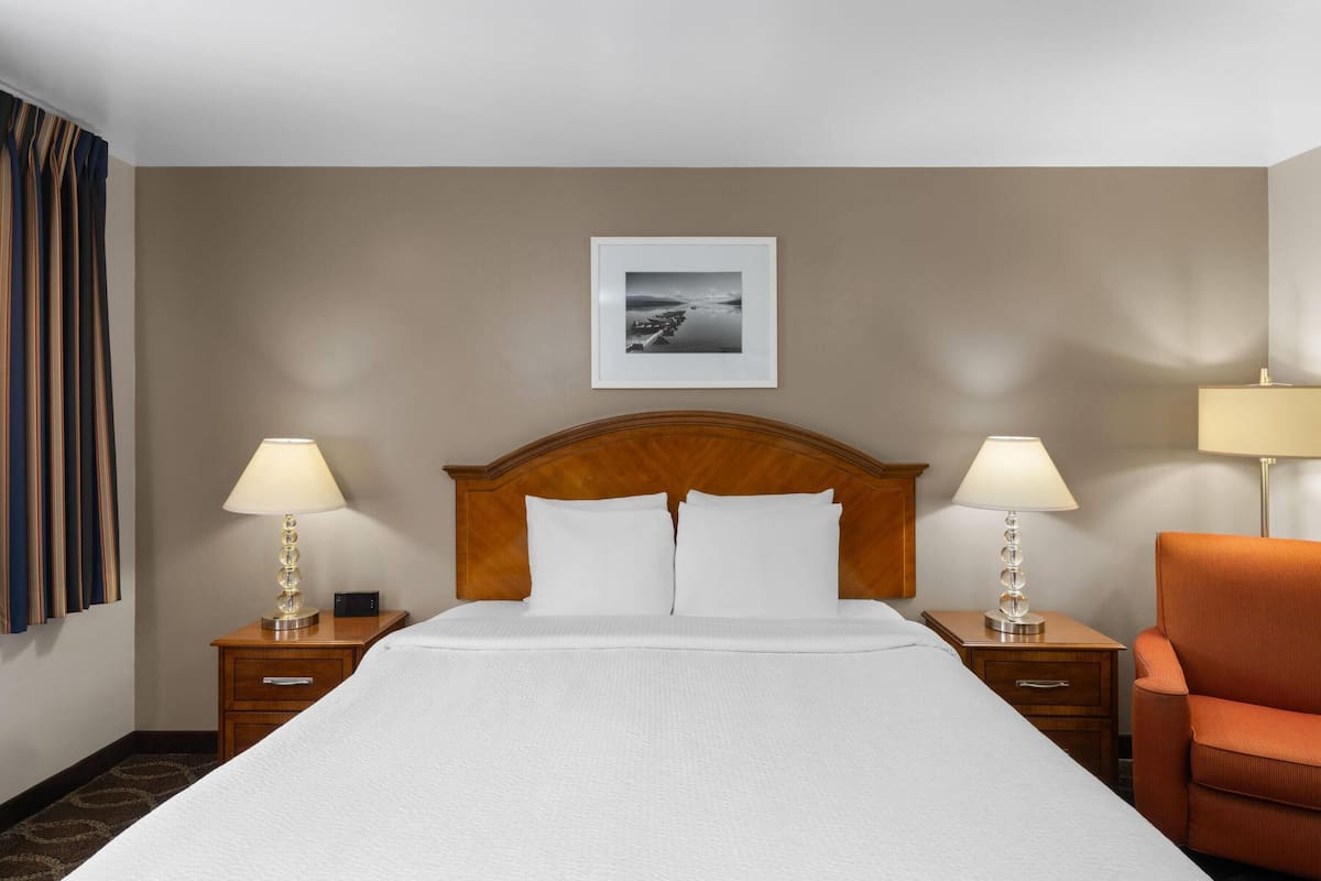 A hotel room with a large bed, two bedside tables with lamps, a wooden headboard, a framed picture on the wall, and an orange chair.