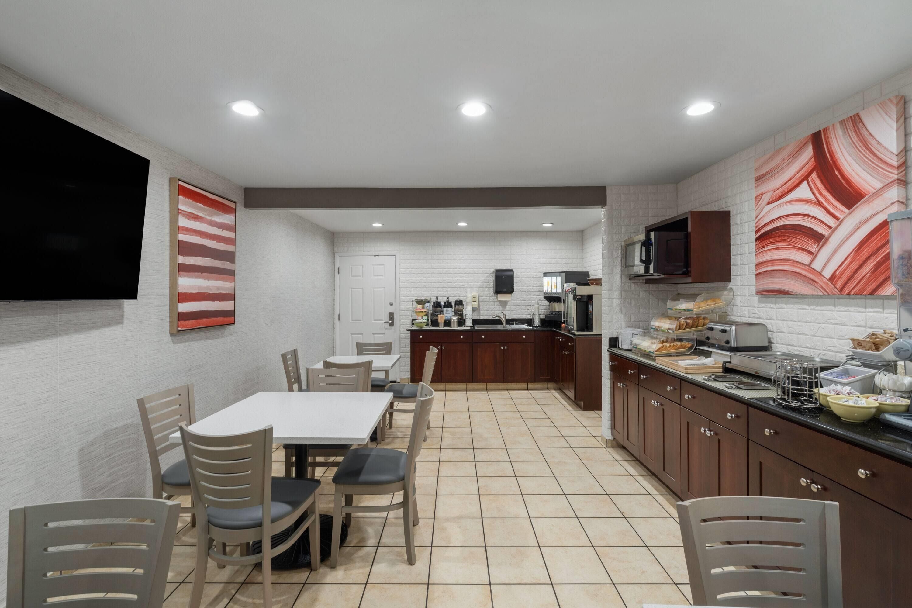 Une cuisine moderne avec des carreaux blancs, des meubles en bois, un coin repas avec une table et des chaises, et un téléviseur fixé au mur.