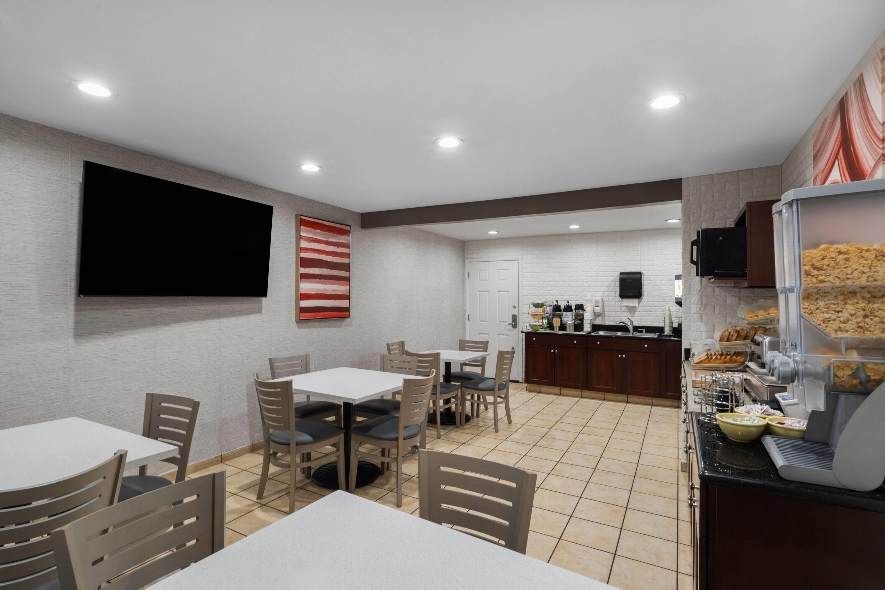 A dining area with tables and chairs, a kitchen area with a coffee machine, and a flat-screen TV mounted on the wall.