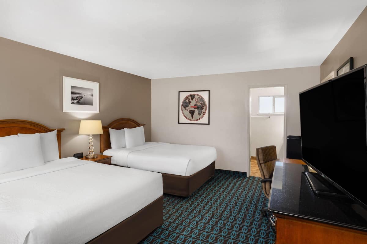 A hotel room with two beds, a desk, a television, and framed pictures on the walls.