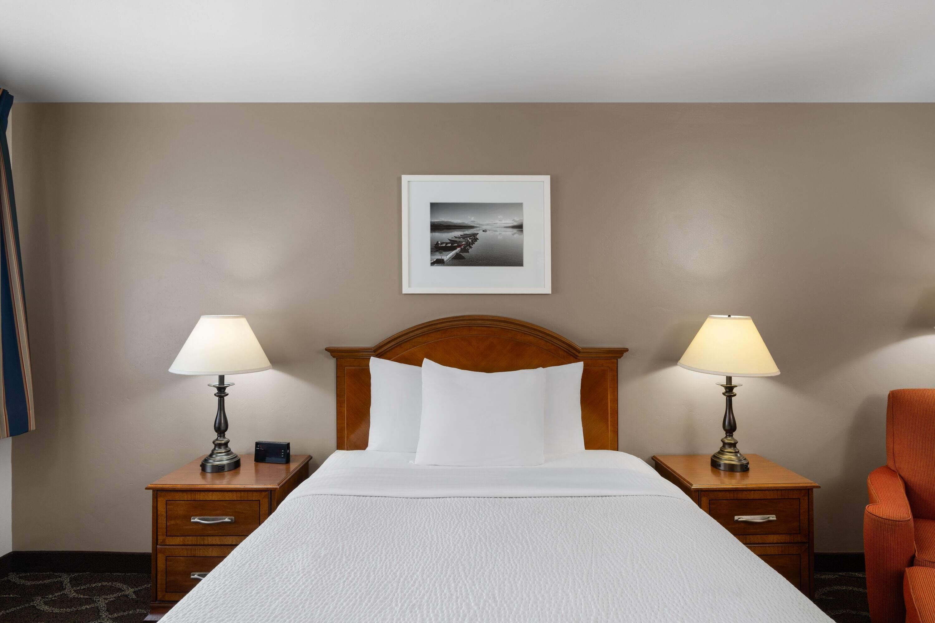 A hotel room with a bed, two bedside tables with lamps, a wooden headboard, a framed picture on the wall, and an orange chair.