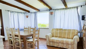 Zona de menjador amb taula i cadires de fusta, un sofà i vista a través de cortines.