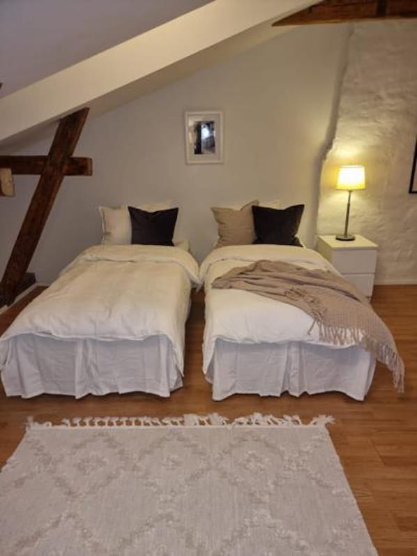 A bedroom with two beds, a wooden ceiling beam, a small bedside table with a lamp, and a patterned rug.