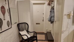 Ein schmaler Flur mit Holzboden, einem schwarzen Stuhl, einem Garderobenhaken mit einem Mantel und einer Tür mit Briefkasten.