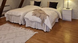 Ein Schlafzimmer mit schrägen Wänden, Holzbalken, zwei Betten mit weißer Bettwäsche, einem Nachttisch mit Lampe und einem Teppich auf dem Boden.
