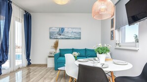 Un salon moderne avec un canapé de couleur turquoise, une table à manger avec des chaises, un tableau représentant des voiliers et une grande fenêtre avec des rideaux.