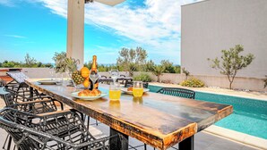 Un espace extérieur aménagé pour les repas, avec une piscine, des chaises en osier et une table dressée avec des assiettes, des verres et un ananas.